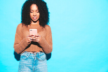 Beautiful black woman with afro curls hairstyle.Smiling model in trendy summer clothes. Sexy carefree female posing near blue wall in studio.Using smartphone apps.Looking at cellphone screen