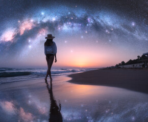 Wall Mural - Arched Milky Way and young woman in hat on sandy beach against starry sky reflected in water at night. Landscape. Summer travel. Girl on the tropical sea coast and bright Milky Way arch, stars. Space