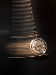 Vintage watch on a chain with a lid swings like a pendulum against a black background and leaves a luminous trail behind it illustrating time flies concept