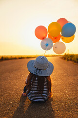 Wall Mural - Little girl sitting on road with colorful balloons in hands at sunset. Back view