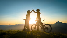 Two Happy Woman High Five Over The Sunset After A Successful Mountain Biking Trip In The Mountains. Celebrate A Cross Country Cycling Journey.