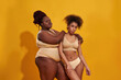 © Svitlana - Two serious african american women in beige underwear with different body size standing together, posing isolated over yellow background