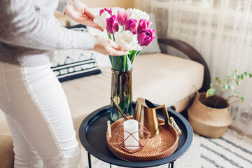 Woman enjoys tulips flowers put in vase on table with tray, watering can and candle. Interior and spring decor at home