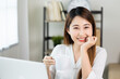 © Chanakon - Young asian beautiful business woman working with laptop sitting at home drinking coffee. Smiling charming happy young female doing homework meeting conference with team at home.