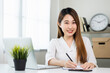 © Chanakon - Young asian beautiful business woman working with laptop sitting at home and note on book. Smiling charming happy young female doing homework meeting conference with team at home.