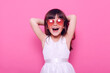 © sementsova321 - Happy small female kid wearing elegant white dress and heart-shaped glasses looks at camera with excited expressing, being glad of great event, raising hands, isolated over pink background.