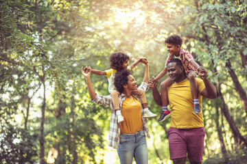 african american family walking trough park. parents carrying children on piggyback.