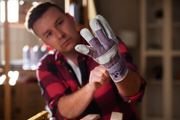 Wall Mural - Young male carpenter working in workshop. Carpenter putting on protective gloves