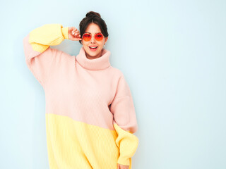 Wall Mural - Young beautiful smiling female in trendy summer colorful sweater. Sexy carefree woman posing near light blue wall in studio. Positive model having fun indoors in sunglasses