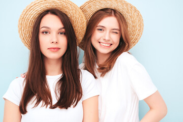 Two young beautiful smiling hipster female in trendy summer white t-shirt and jeans clothes.Sexy carefree women posing near light blue wall in studio.Positive models in hats