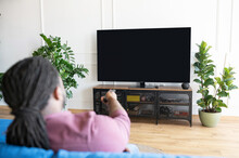 Back View African-American Guy With Dreadlocks Holds Remote Controller And Switching Tv Channels, Watching TV Sitting On The Couch At Home, The Lcd TV With Empty Black Screen, Copy Space, Mockup