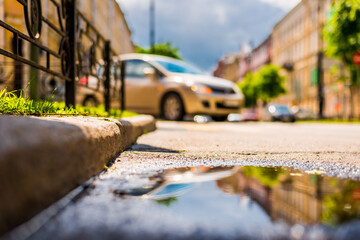 Naklejka na meble Sunny day after rain in the city, the car turns from the corner of the road. Close up view from a puddle level near a curb
