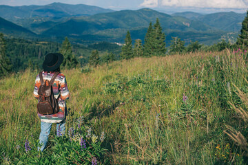 Wall Mural - Woman traveler with backpack and hat standing on beautiful hill and looking on mountains. Wanderlust