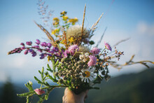 Beautiful Colorful Bouquet Of Wildflowers In Hand On Background Of Mountain Hills And Sunny Sky