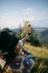 Wall Mural - Woman traveler in hat holding bouquet of wildflowers on background of mountain hills and sky. Travel