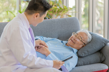 An old fat asian male patient wearing light blue long sleeve shirt and eyeglasses lay down on gray pillows and  sofa smiling and talking to young male doctor wearing white lab coat sitting on sofa 