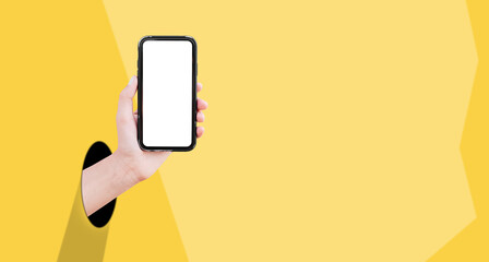 Close-up of male hand coming out from the hole in paper, holding smartphone with mockup on background of yellow color.