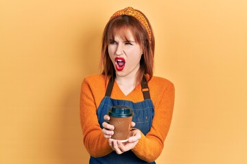 Canvas Print - Redhead young woman wearing waitress apron holding take away cup of coffee angry and mad screaming frustrated and furious, shouting with anger. rage and aggressive concept.