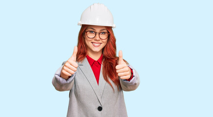 Wall Mural - Young redhead woman wearing architect hardhat approving doing positive gesture with hand, thumbs up smiling and happy for success. winner gesture.