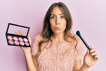 Wall Mural - Young brunette woman holding makeup brush and blush puffing cheeks with funny face. mouth inflated with air, catching air.