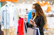 © maxbelchenko - Fashionable  woman in protective medical mask with multi-colored  bags in modern shop center. Young woman using her phone. The joy of consumption, Gift shopping, holiday. Online shopping concept.
