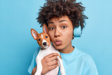Lovely African American Woman Carries Small Breed Dog Near Face Keeps Lips Folded As If Wanting To Kiss Someone Loves Her Favorite Pet Have Walk Dressed Casually Listens Music In Stereo Headphones