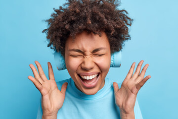 Wall Mural - Overjoyed Afro American woman keeps hands raised exclaims joyfully closes eyes from happiness reacts on awesome news wears wireless headphones on ears isolated over blue background. Joy concept