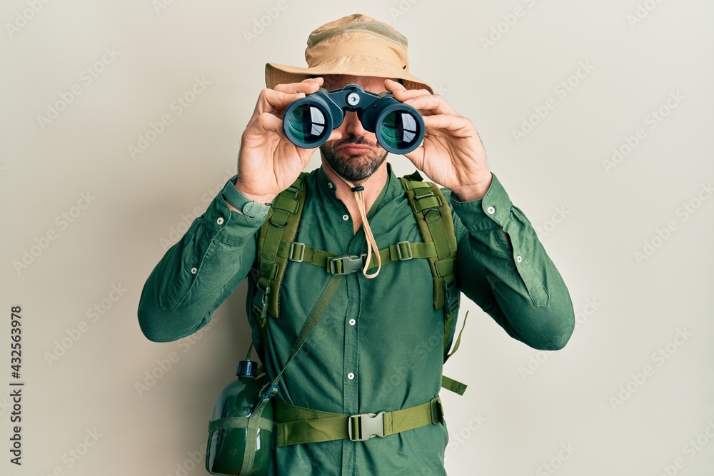 Handsome man with beard wearing explorer hat looking through binoculars skeptic and nervous, frowning upset because of problem. negative person. - obrazy, fototapety, plakaty 