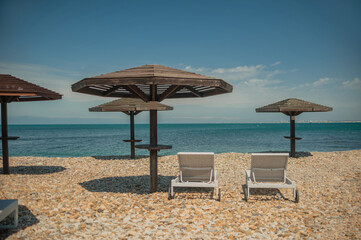 Wall Mural - two white sun loungers under a wooden beach umbrella on a rocky beach in summer on a clear day without people