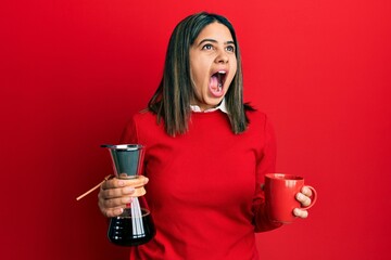 Poster - Young latin woman holding coffee filter and cup angry and mad screaming frustrated and furious, shouting with anger looking up.