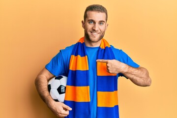 Young caucasian man football hooligan cheering game holding ball pointing finger to one self smiling happy and proud