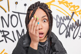Fototapeta Młodzieżowe - Startled teenage girl covers face with hand stares surprised at camera has colorful nails braided hairstyle reacts on something amazing poses against graffiti background. Youth lifestyle urban style