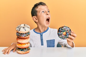 Wall Mural - Adorable caucasian kid eating doughnuts for breakfast angry and mad screaming frustrated and furious, shouting with anger. rage and aggressive concept.