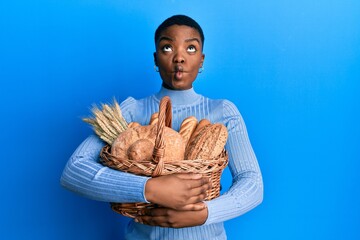 Sticker - Young african american woman holding wicker basket with bread making fish face with mouth and squinting eyes, crazy and comical.