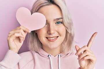 Sticker - Young blonde girl holding heart shape paper over eye smiling happy pointing with hand and finger to the side