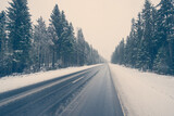 Fototapeta Na ścianę - Empty winter road during a snowfall passing through a spruce forest. View from the road, image in the blue toning