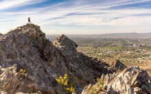 A Lone Hiker Is Seen Atop Piestewa Peak In Phoenix, Arizona, On Top Of A Rocky Cliff Overlooking The Valley Of The Sun With A Blue Sky With High Clouds. 