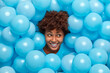 Glad brunette young African American woman with curly hair sticks out head through inflated blue balloons smiles broadly has happy mood celebrates something. Holiday decor. Festive atmosphere.