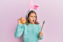 Little Beautiful Girl Wearing Cute Easter Bunny Ears Holding Colored Egg Angry And Mad Screaming Frustrated And Furious, Shouting With Anger. Rage And Aggressive Concept.