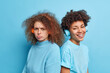 © WHstudio Leushin N - Indoor shot of displeased curly haired woman and her best friend companion stand back to each other listen music in wireless headphones isolated over blue background. Diverse women pose closely
