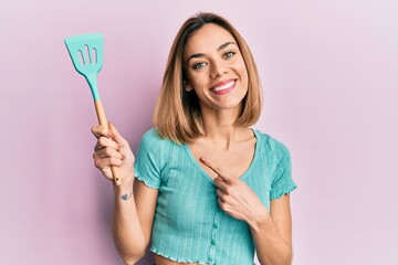 Young caucasian blonde woman holding silicone spatula smiling happy pointing with hand and finger