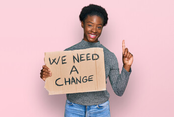 Poster - Young african american girl holding we need a change banner surprised with an idea or question pointing finger with happy face, number one