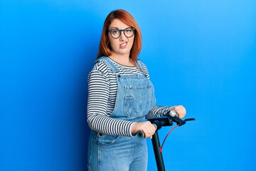 Canvas Print - Beautiful redhead woman standing on modern scooter over blue background clueless and confused expression. doubt concept.