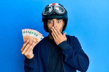 Canvas Print - Young hispanic man wearing motorcycle helmet holding 10 euros covering mouth with hand, shocked and afraid for mistake. surprised expression