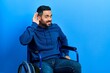 Handsome hispanic man with beard sitting on wheelchair smiling with hand over ear listening an hearing to rumor or gossip. deafness concept.