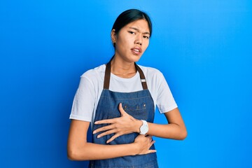 Poster - Young chinese woman wearing waiter apron with hand on stomach because nausea, painful disease feeling unwell. ache concept.