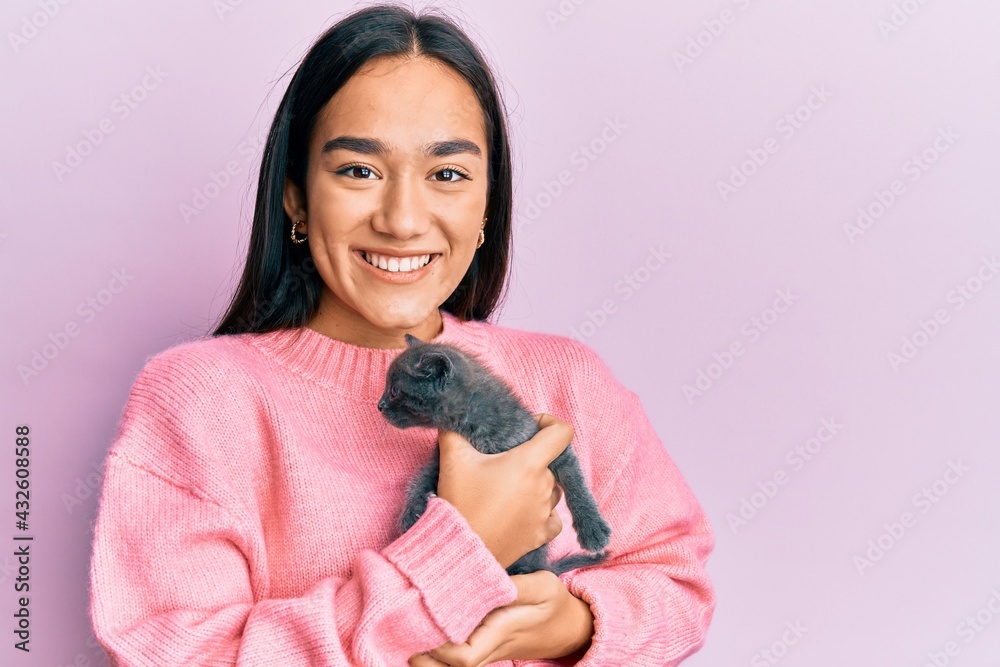 Young hispanic girl smiling happy holding cute cat over isolated pink background.