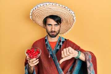 Young hispanic man wearing mexican hat holding chili with angry face, negative sign showing dislike with thumbs down, rejection concept