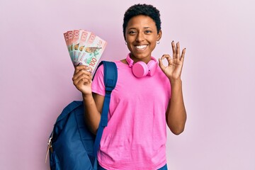 Wall Mural - Young african american girl wearing student backpack and holding new zealand dollars doing ok sign with fingers, smiling friendly gesturing excellent symbol