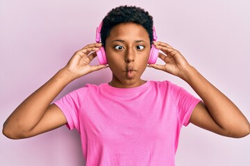 Wall Mural - Young african american girl listening to music using headphones making fish face with mouth and squinting eyes, crazy and comical.